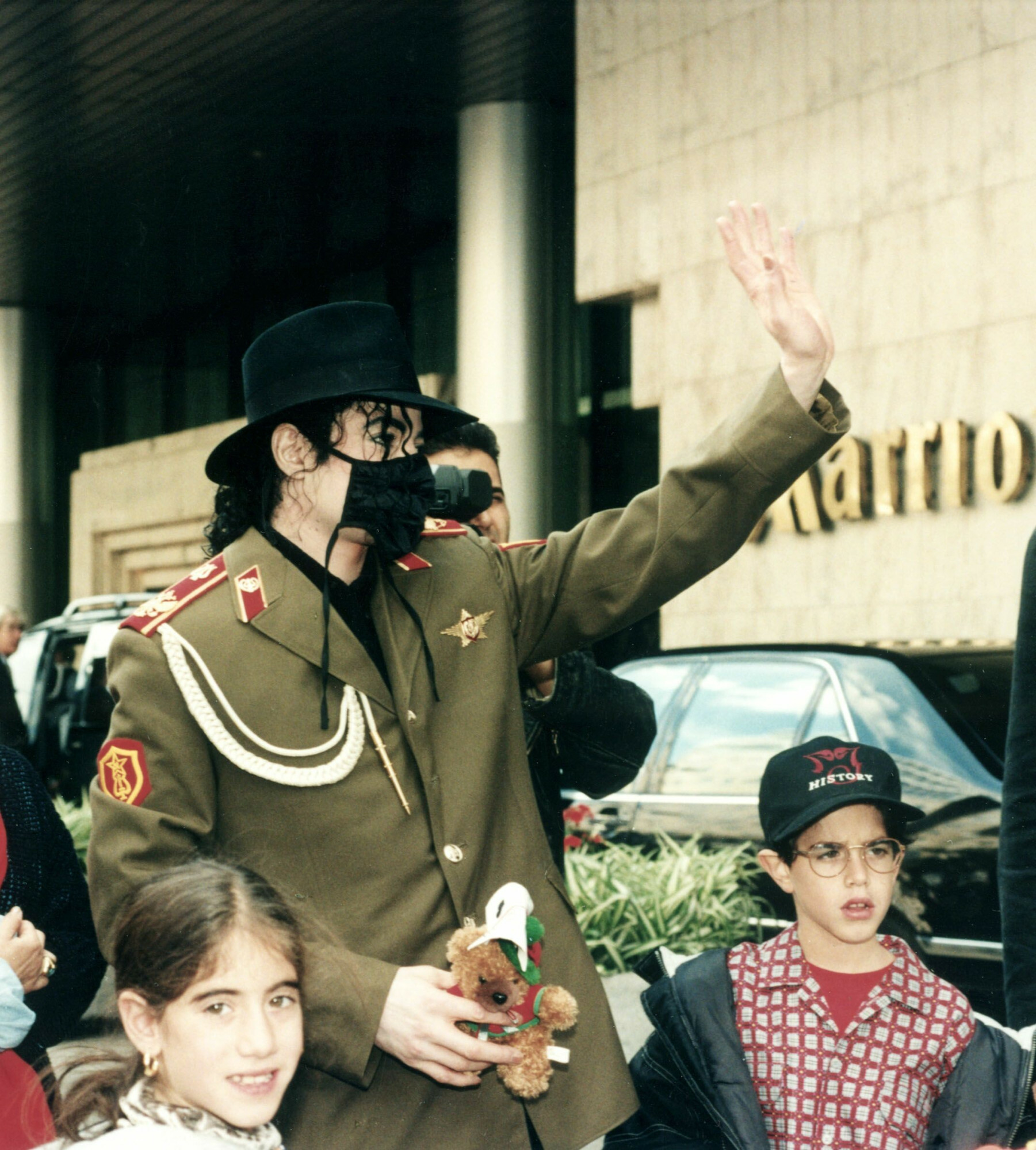 Michael Jackson w oliwkowym mundurze i czarnej maseczce macha do fanów przed hotelem Marriott, obok dzieci z maskotką